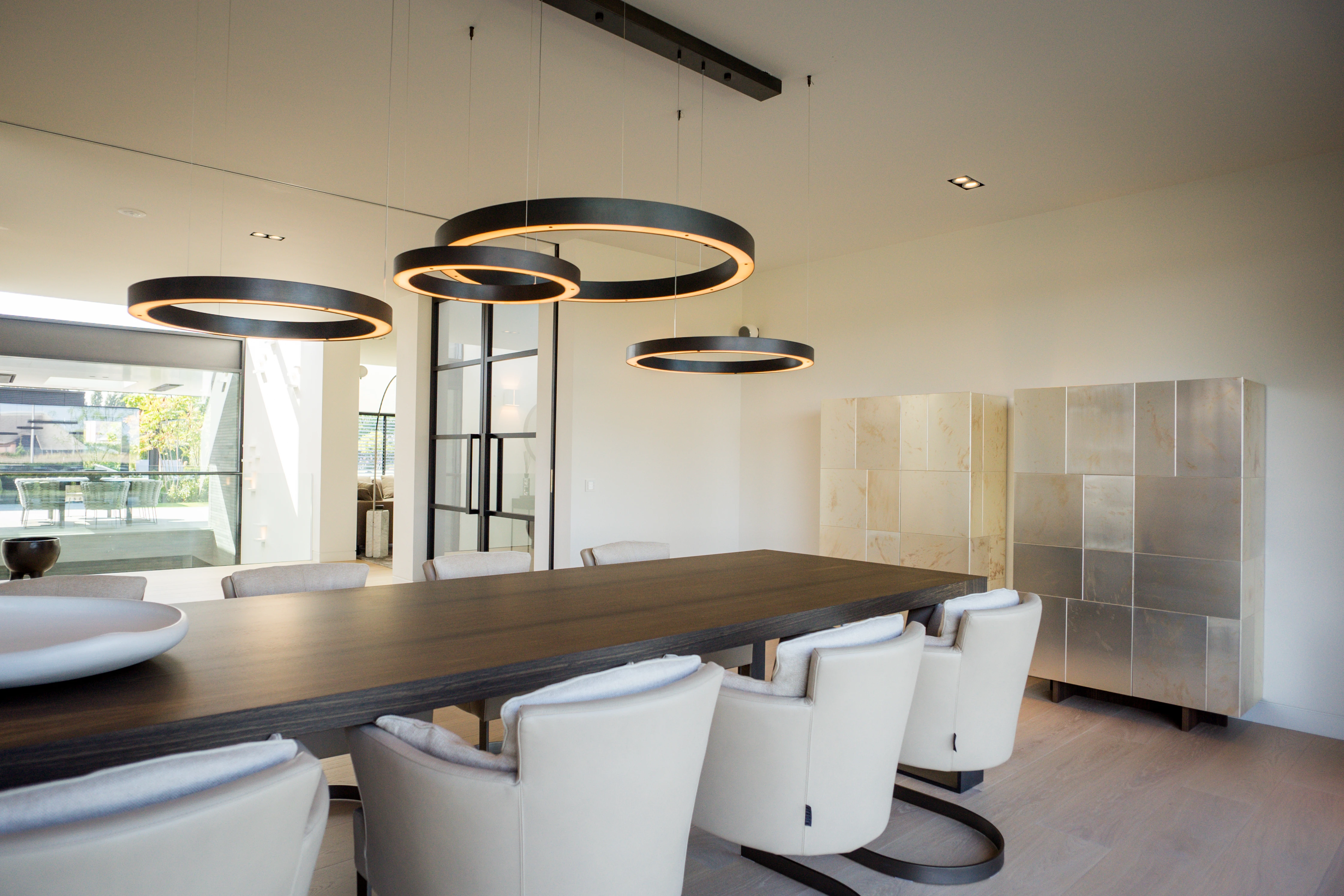Eetkamer met een lange donkere tafel en drie ronde lichtringen boven de tafel