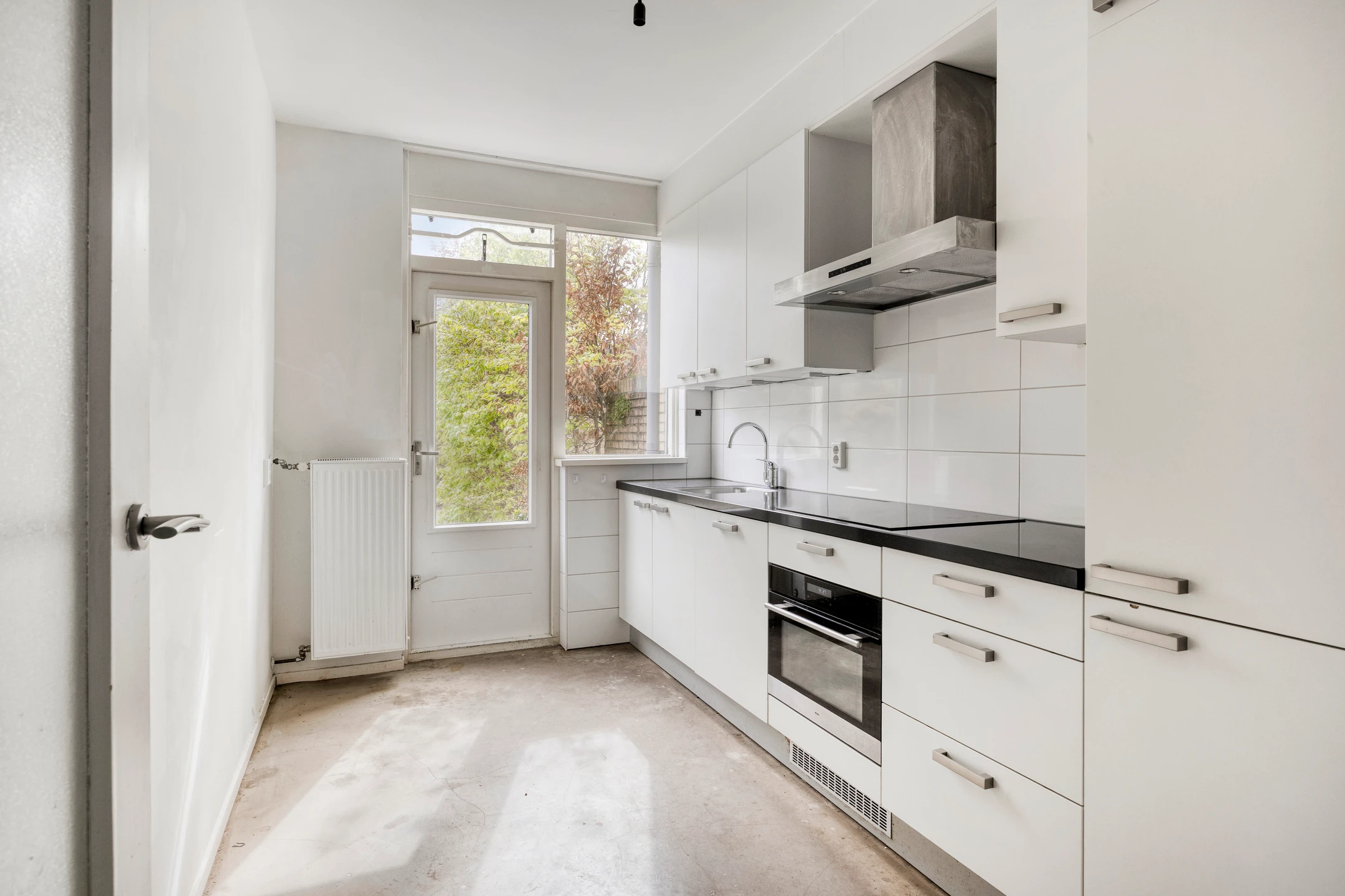 Smalle keuken met witte kasten, een zwart aanrechtblad, een witte tegelwand en een kale betonvloer