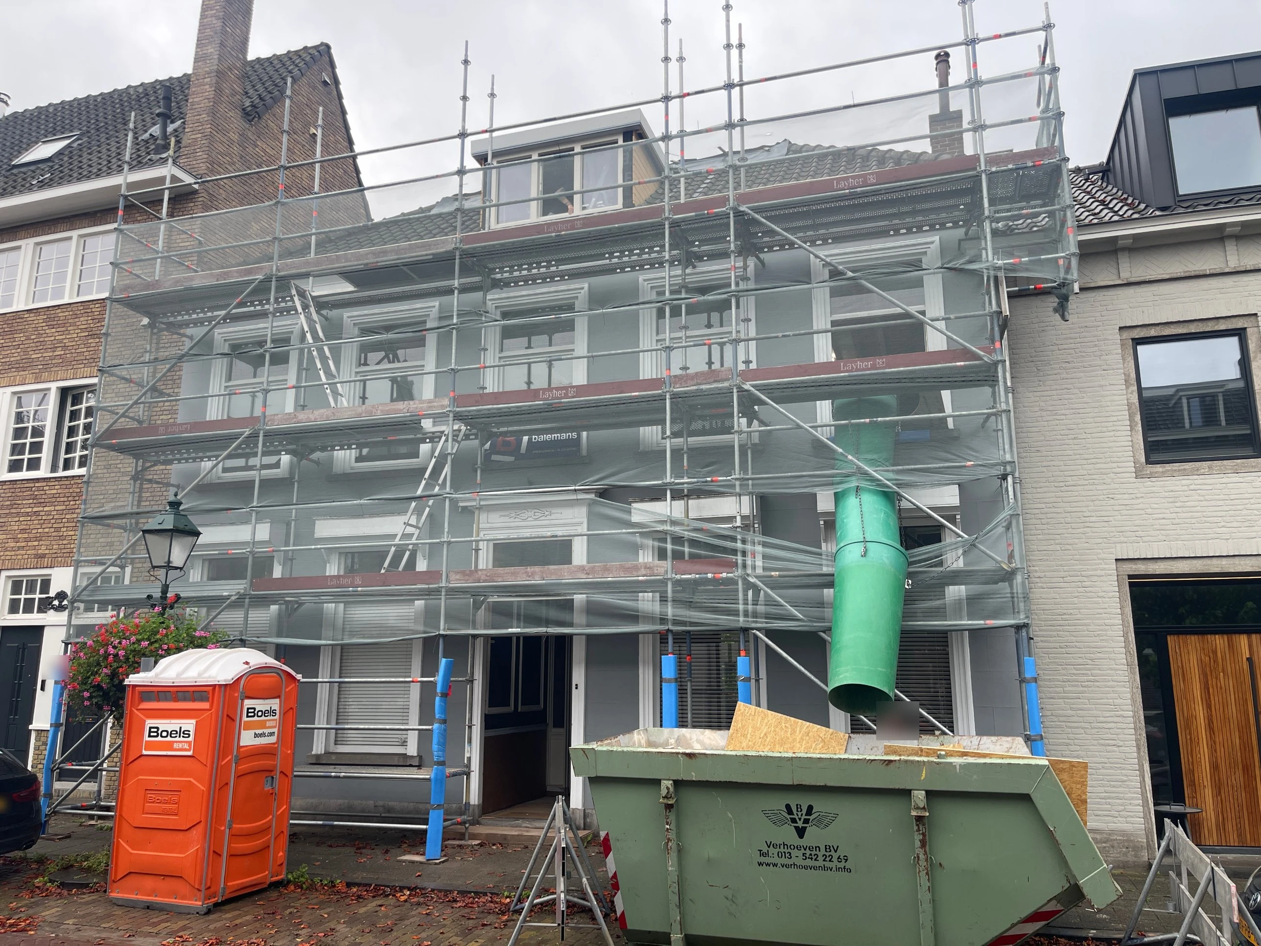 Voorgevel van de woning volledig in de steigers, met een stortkoker voor bouwafval en een container ervoor