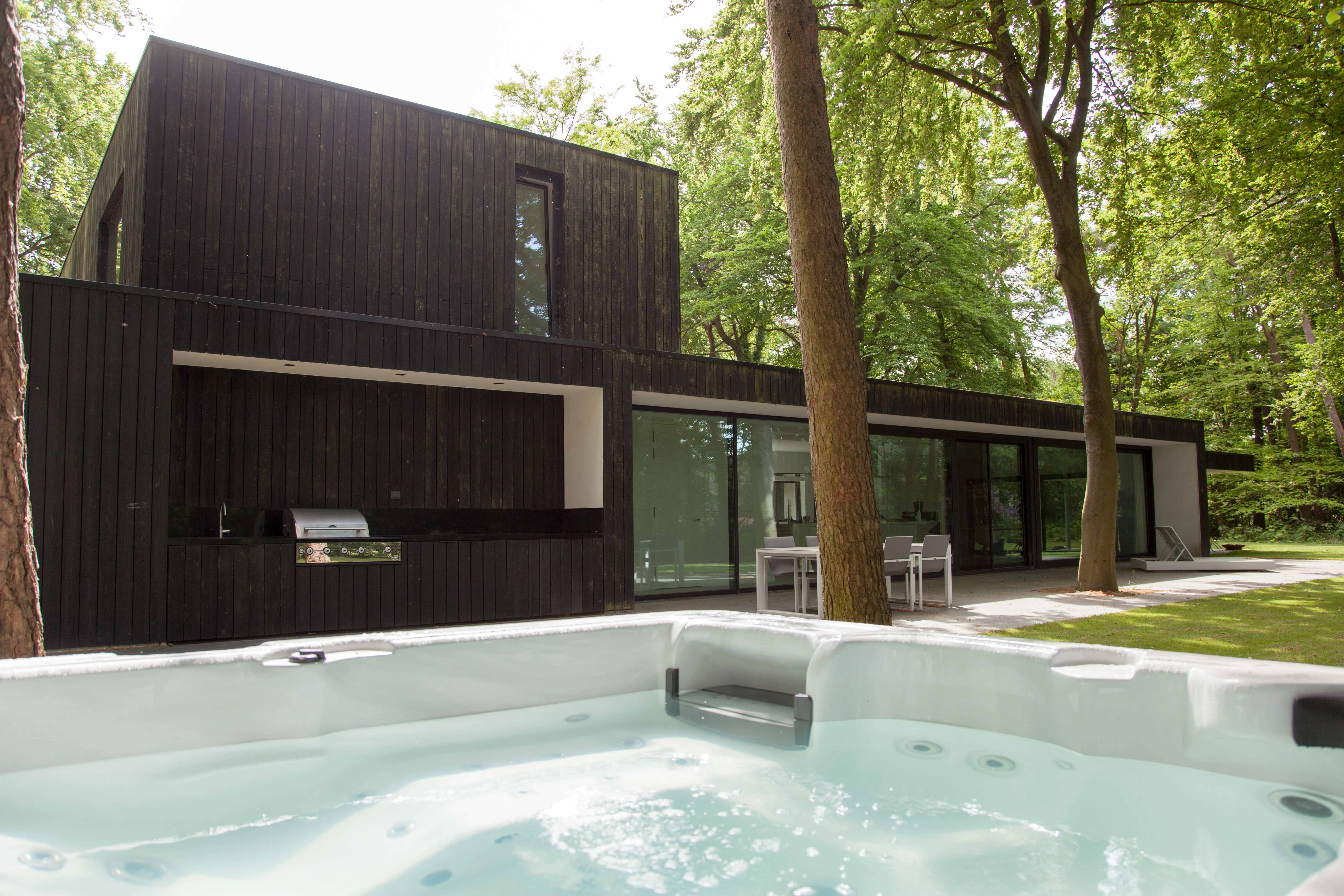 Achtergevel met zwart gevelhout, een verzonken whirlpool op de voorgrond en een buitenkeuken en eettafel onder de overkapping