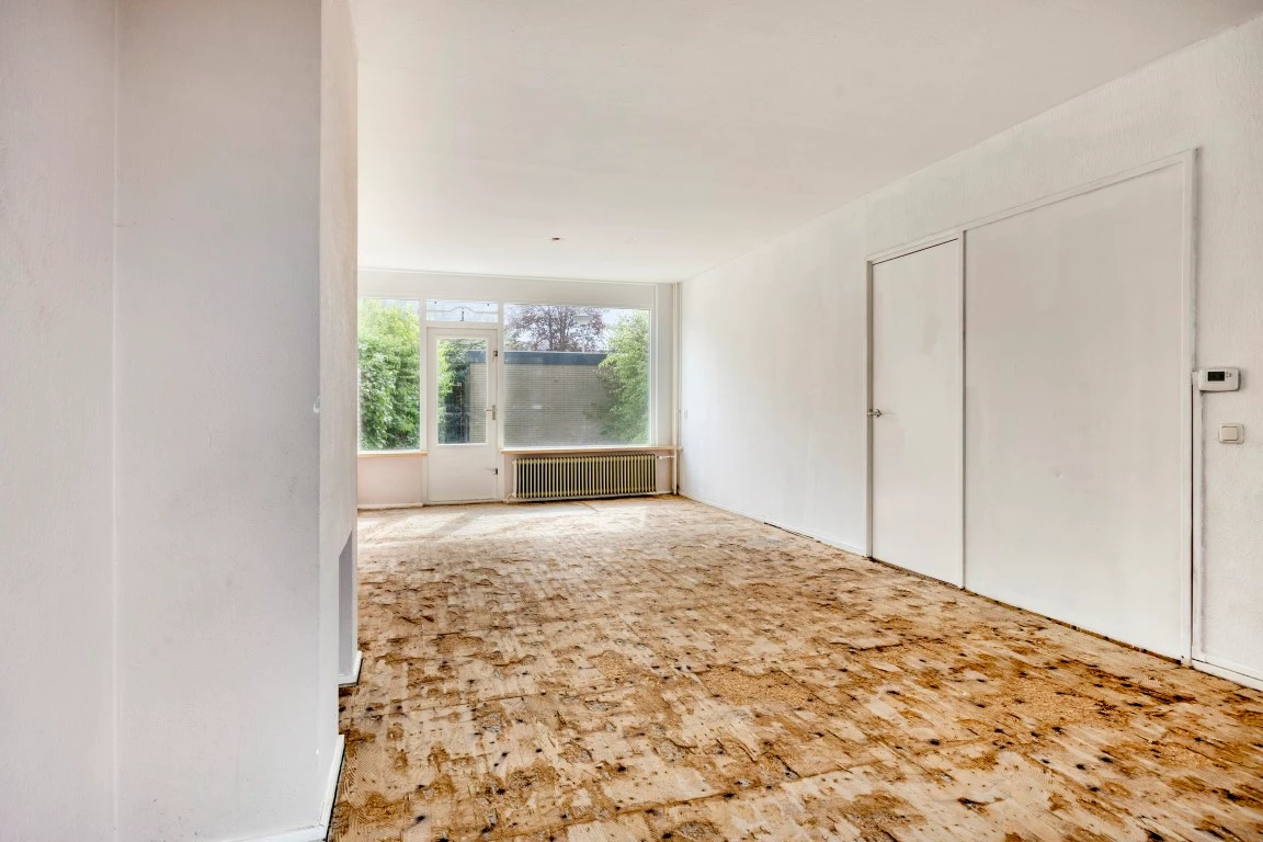Lege woonkamer met witte wanden en een kale houten ondervloer, met uitzicht op de tuin